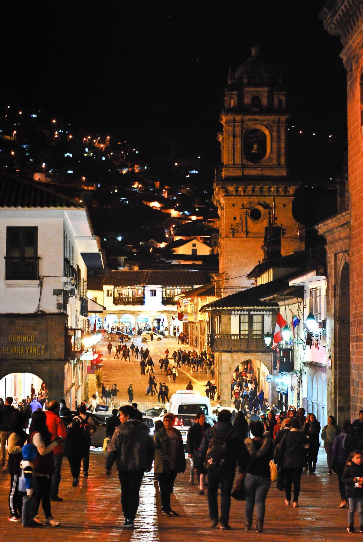 Nighttime street scene in a historic colonial town with crowds, illuminated church tower, and white buildings on hillside