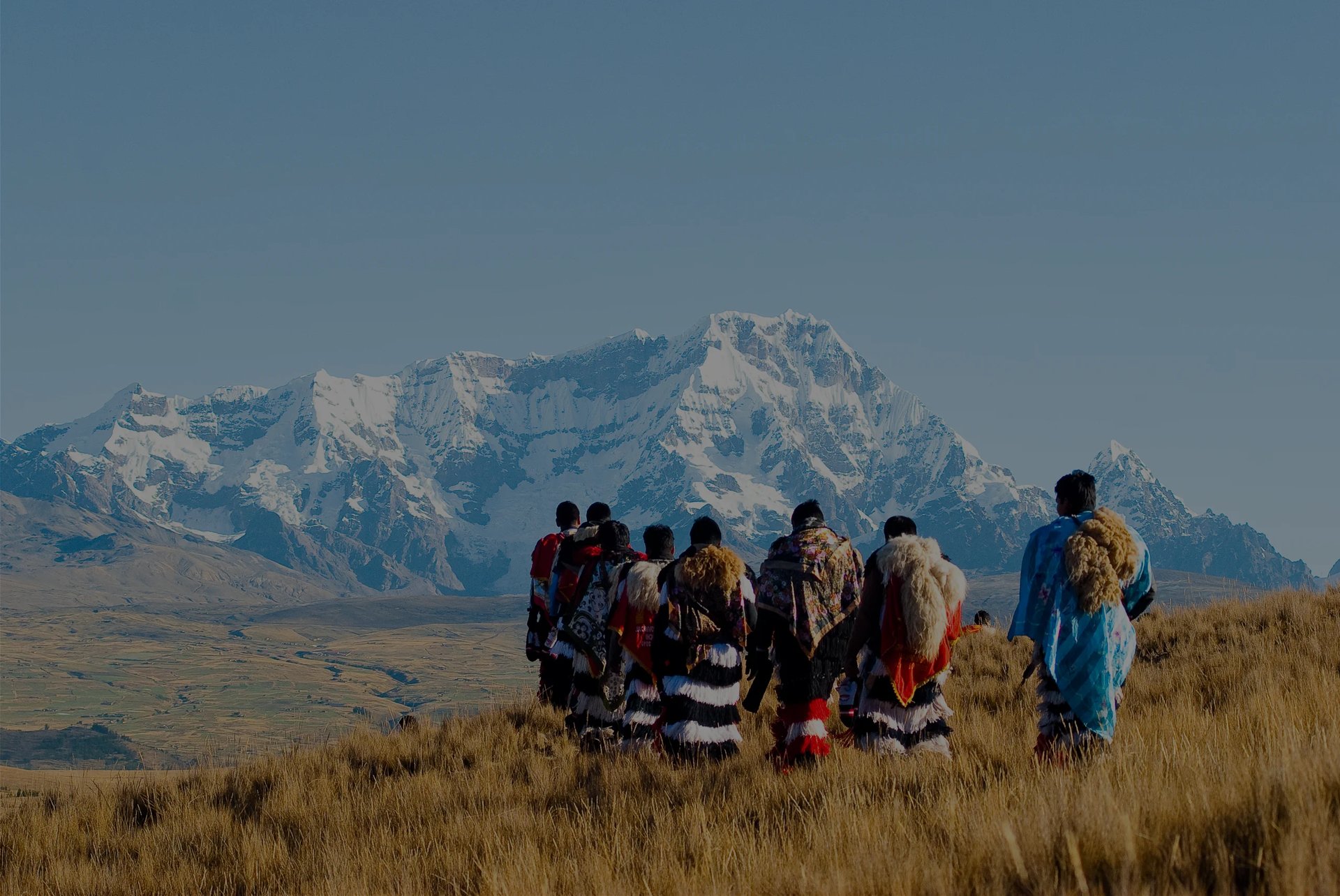Group of people in traditional clothing standing in grassland with snow-capped mountains in the background