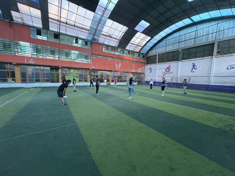 Indoor soccer field with green turf, several players on field, red brick building walls, and curved glass ceiling with overhead lighting