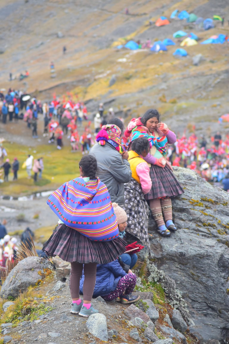 Indigenous women and children in colorful traditional clothing standing on a mountain rock with tents and crowds visible in the valley below