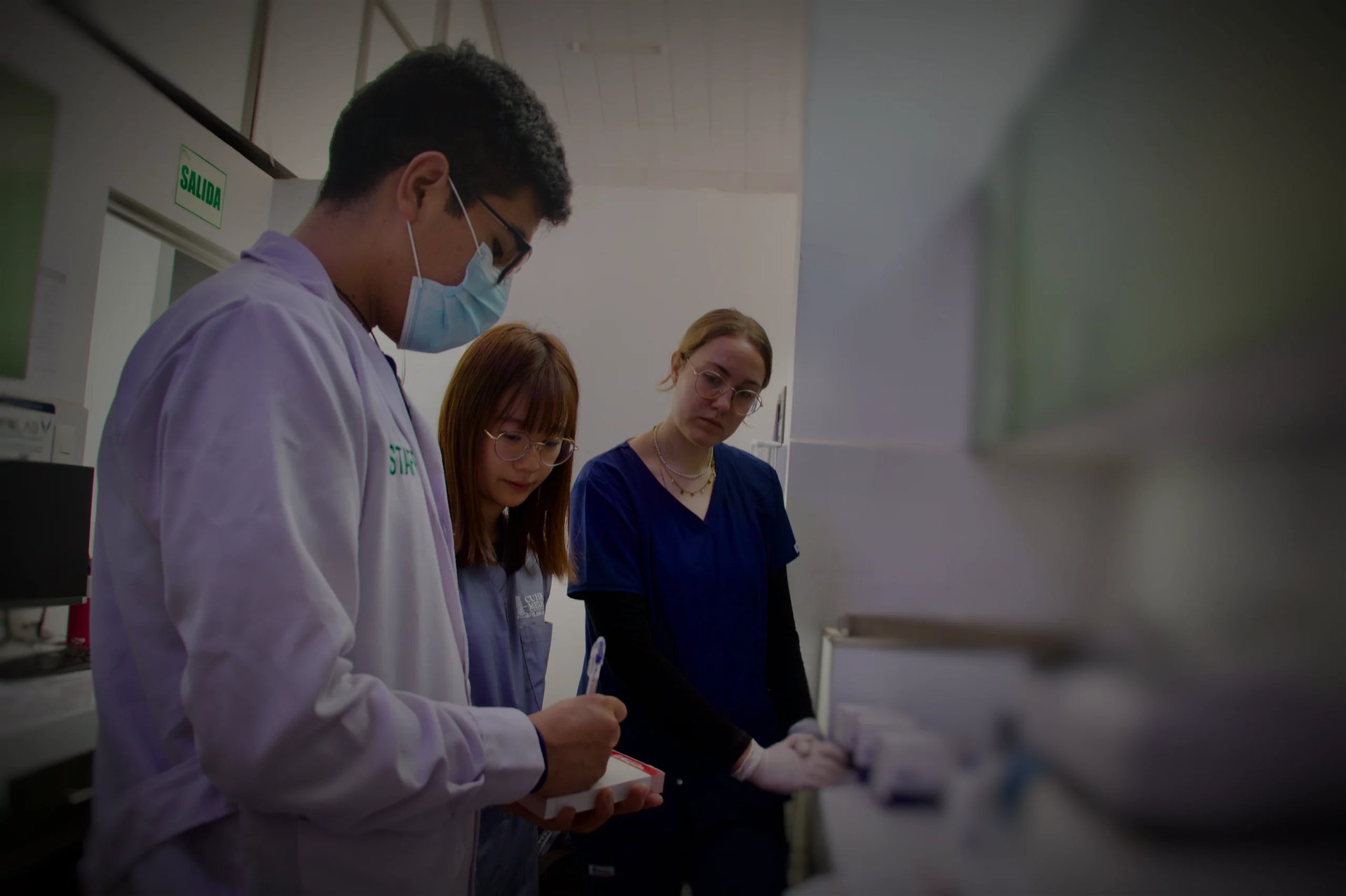 Healthcare worker in purple scrubs and mask instructs two women in a clinical laboratory setting