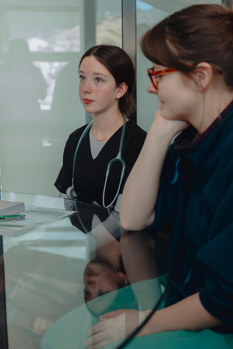 Two healthcare professionals in scrubs having a discussion at a desk with a glass partition in background