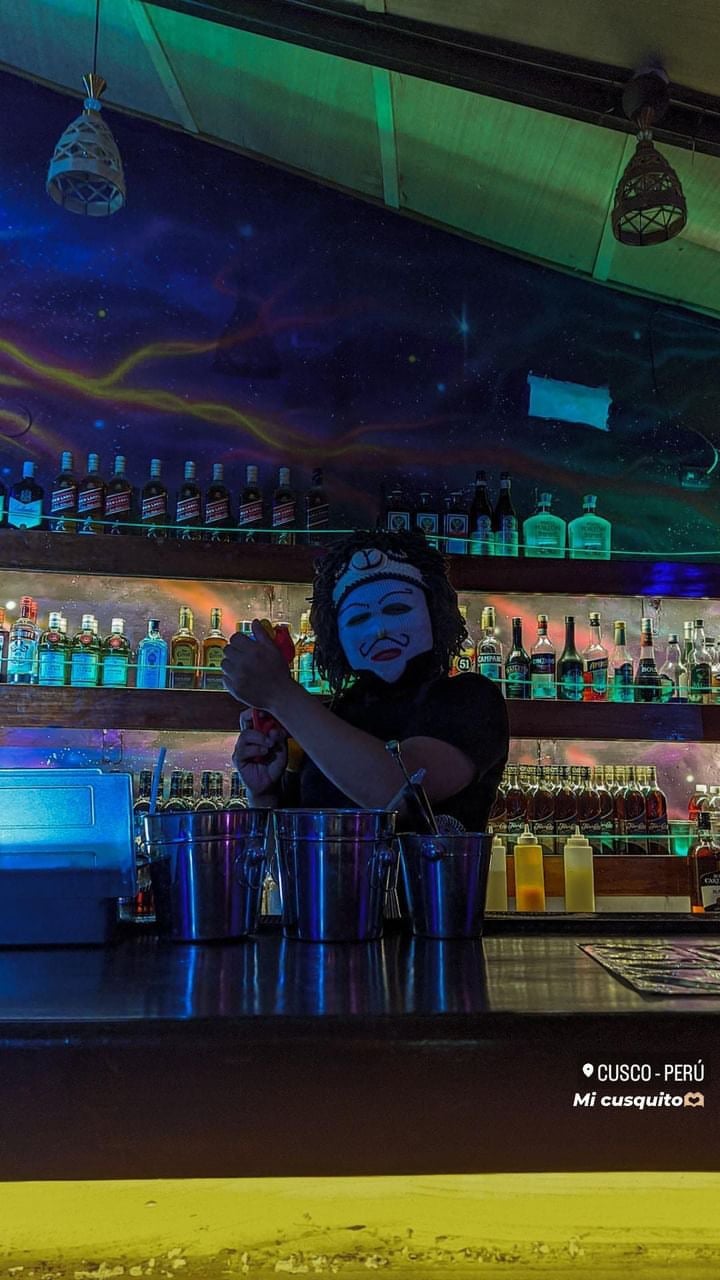 Neon-lit bar interior in Cusco, Peru with illuminated blue face sculpture, hanging decorative lanterns, and shelves of colorful liquor bottles