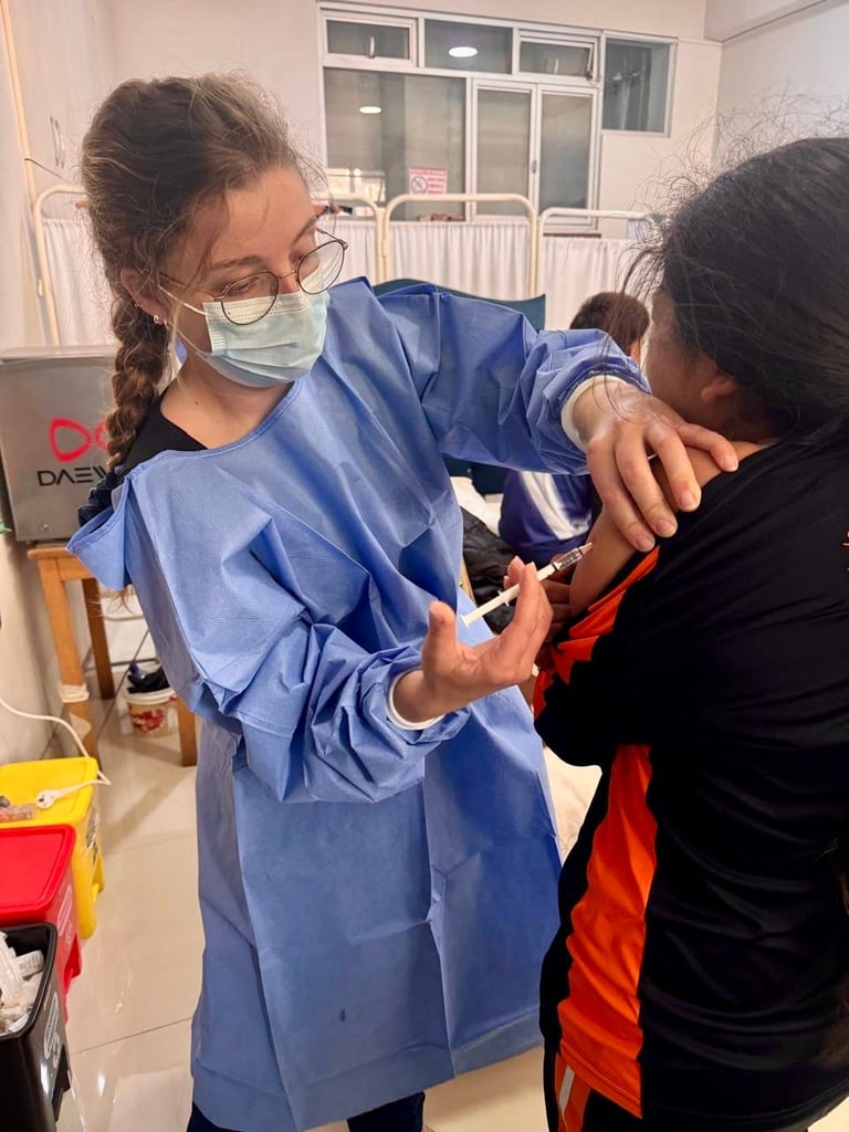 Healthcare worker in blue gown and mask helps another person put on protective equipment in a medical clinic setting