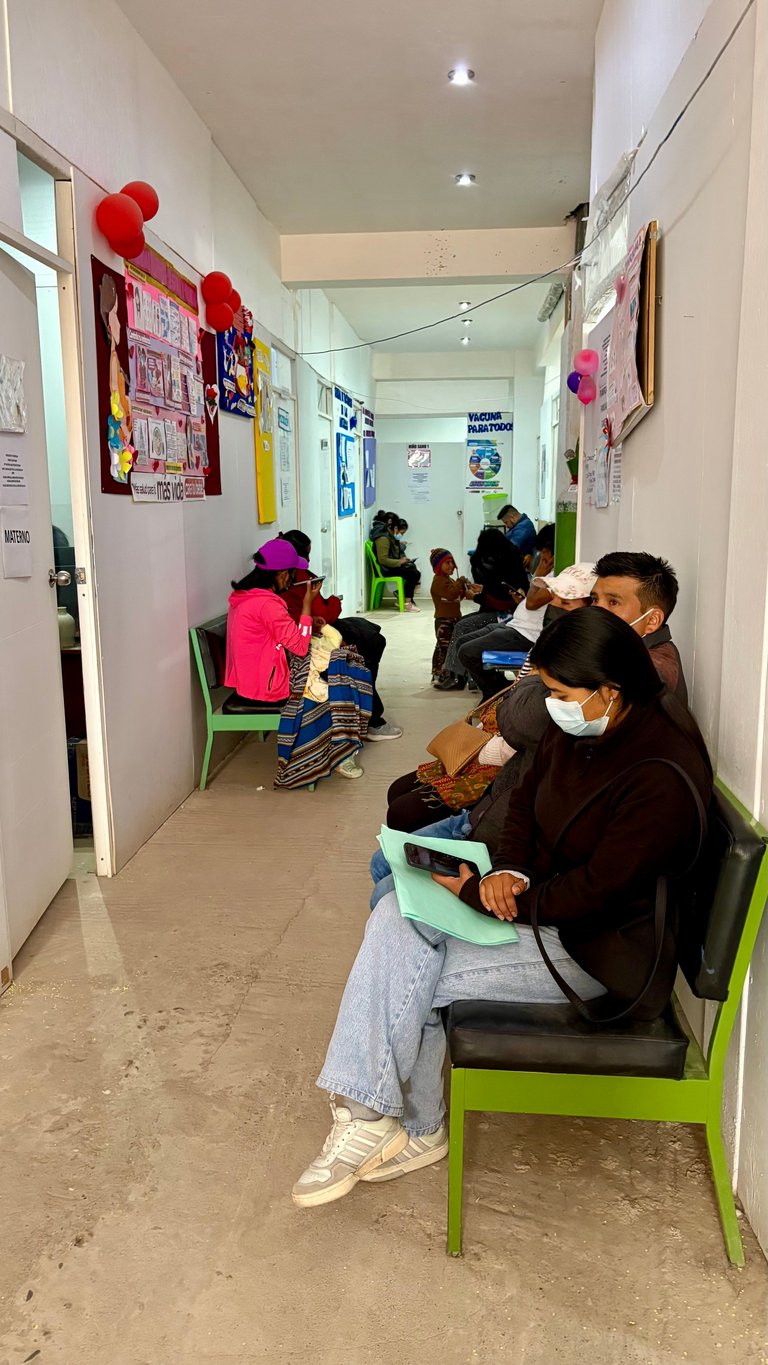 People waiting in a narrow hallway lined with green chairs and educational posters on white walls