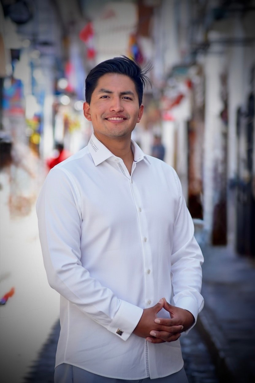 A smiling man wearing a white button-up shirt stands in a blurred outdoor street setting with colorful decorations in the background.