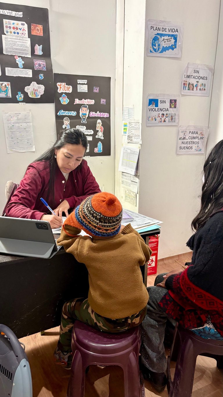 Woman in burgundy jacket tutoring a young child wearing a knit hat and mustard sweater at a desk in a classroom with educational posters on walls